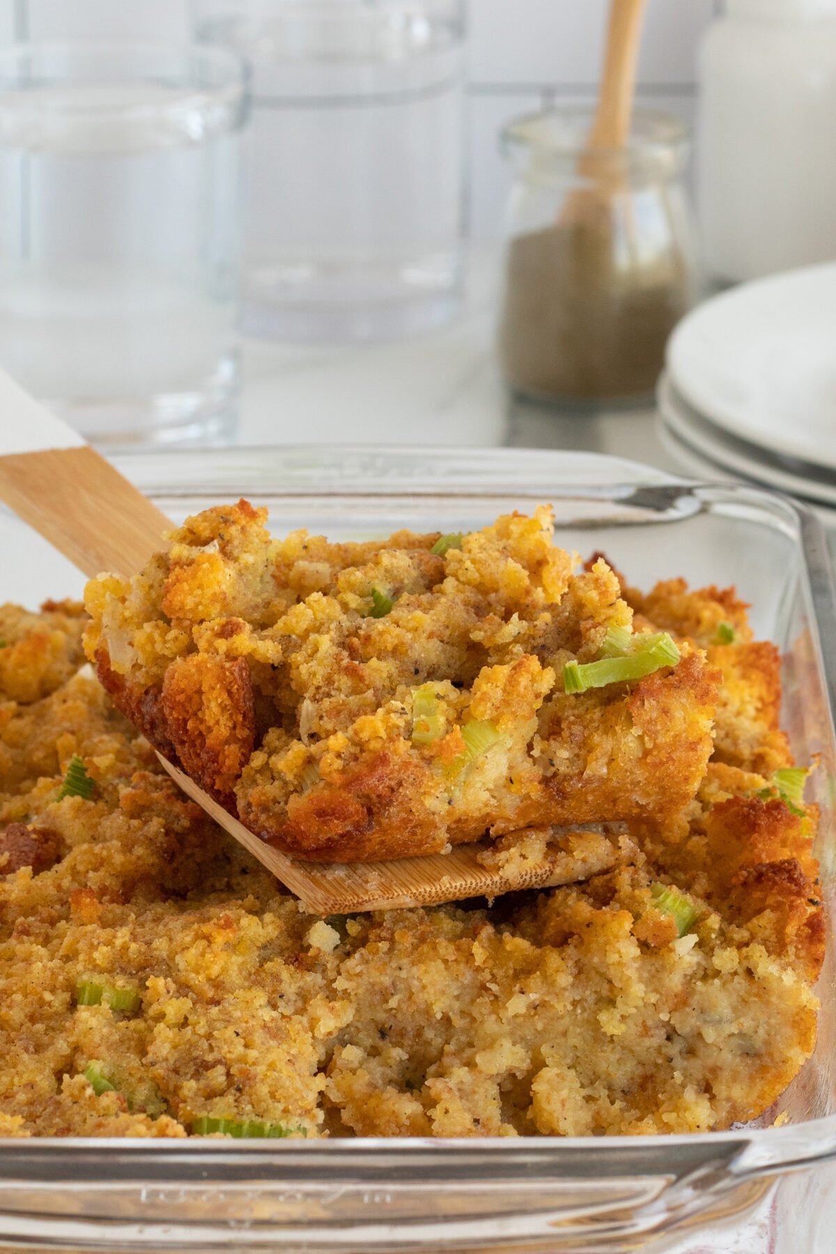 A wooden spatula lifts a portion of golden-brown cornbread stuffing with visible celery pieces from a glass baking dish. Plates and glasses are blurred in the background.