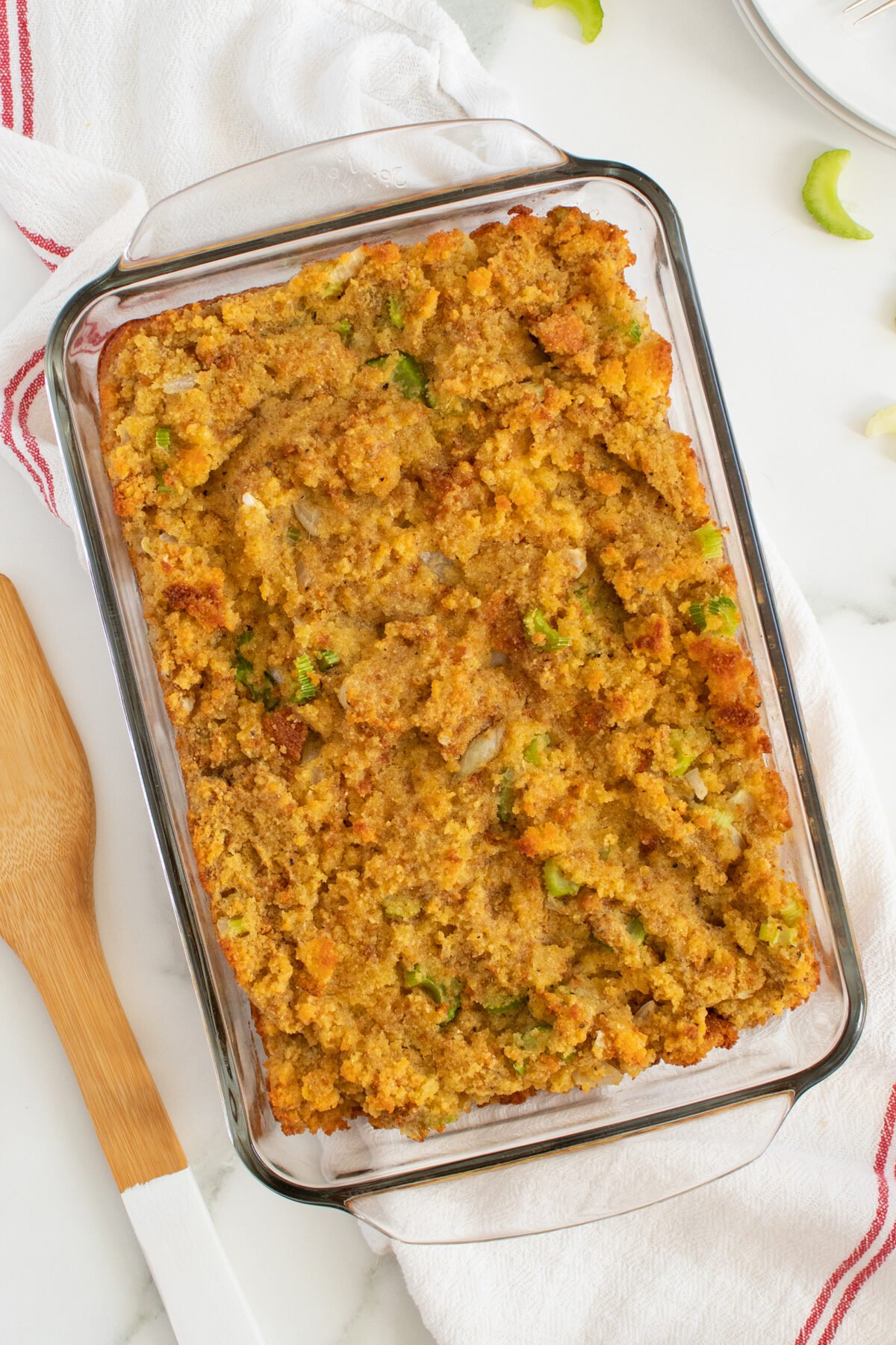 A glass baking dish filled with golden brown cornbread stuffing mixed with celery and seasonings, resting on a white cloth next to a wooden spatula and a striped kitchen towel.