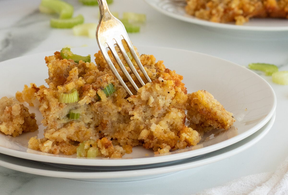 A close-up of a serving of bread stuffing with chopped celery on a white plate. A fork is inserted into the stuffing, and another plate with stuffing is blurred in the background.