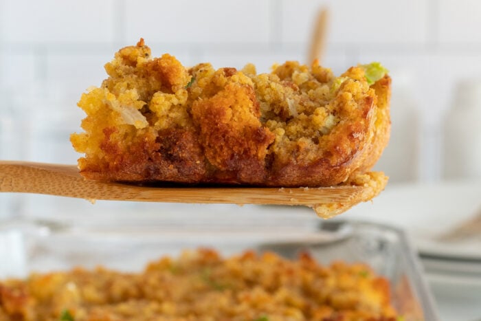 A close-up of a wooden spoon holding a serving of golden brown cornbread stuffing, with a glass baking dish of more stuffing in the background.