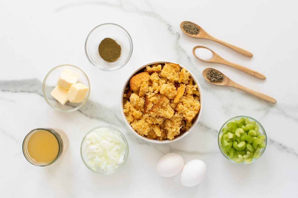 Bowls of crumbled cornbread, chopped celery, diced onion, butter, ground sage, black pepper, and salt, plus two eggs and a small glass dish of broth on a marble countertop.