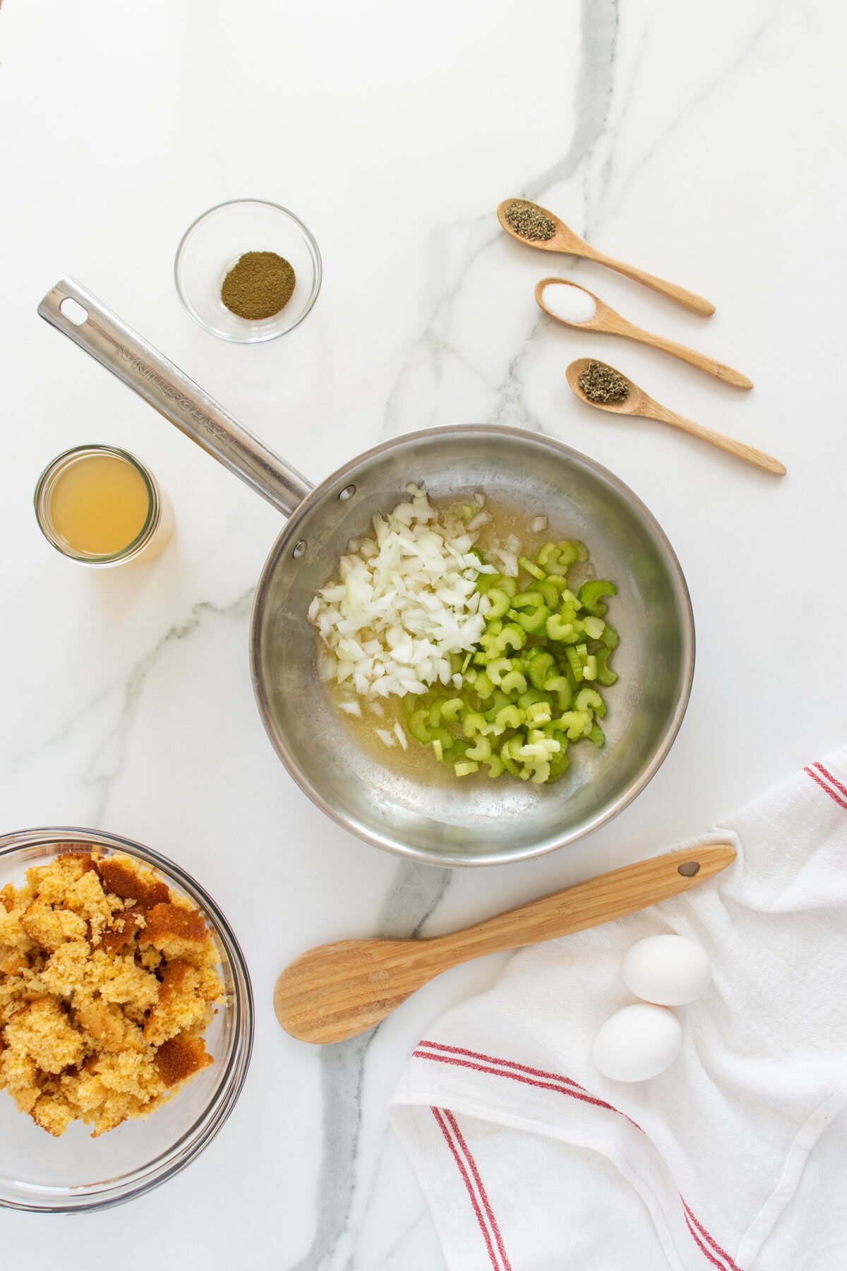 A saucepan with chopped onions and celery, surrounded by a bowl of crumbled cornbread, two eggs, a wooden spoon, small bowls of broth and sage, and three measuring spoons with salt and pepper on a white countertop.