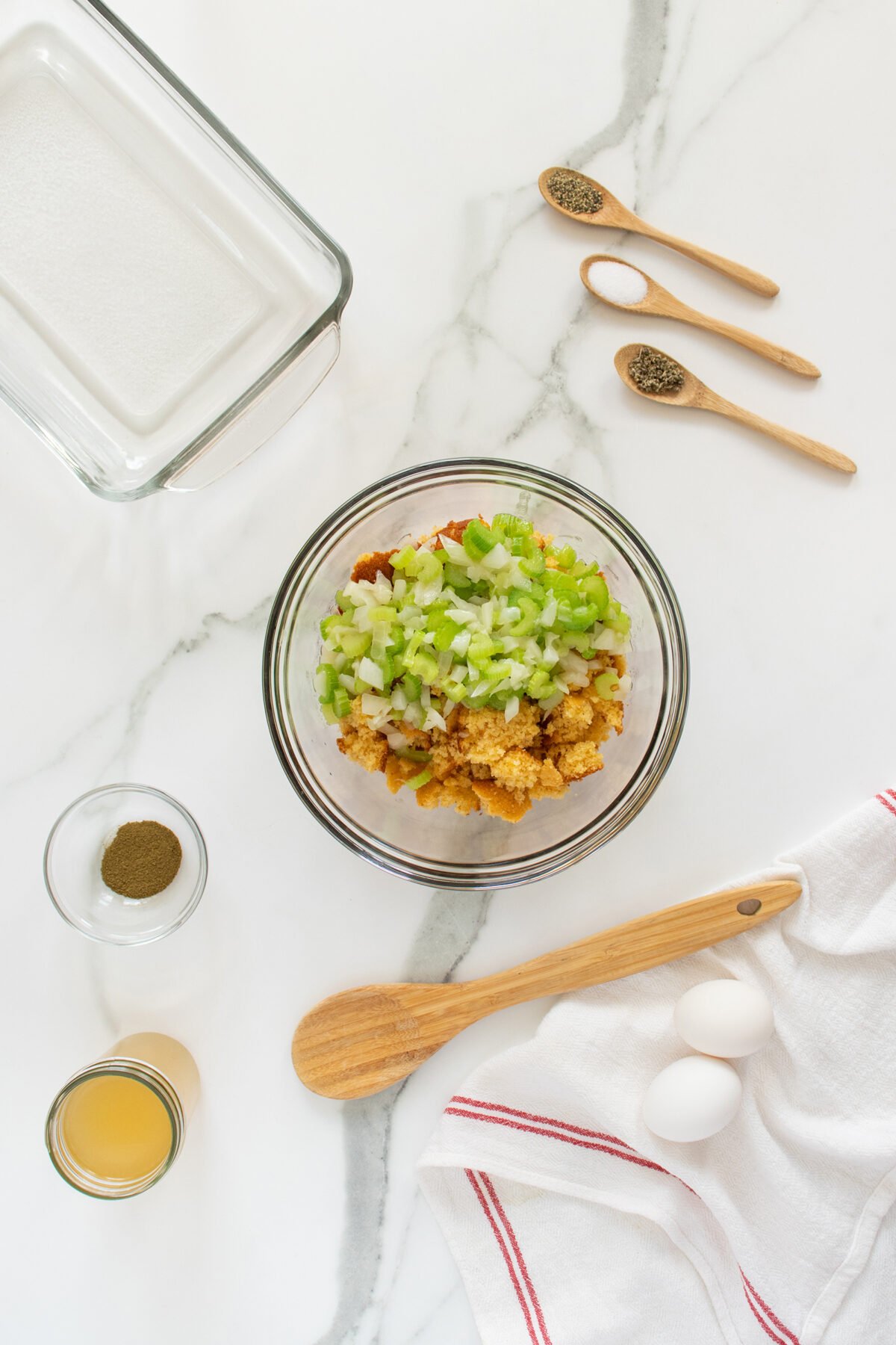 A glass bowl with cornbread cubes, chopped celery, and onions sits on a white marble surface. Nearby are eggs, spices in wooden spoons, a small bowl of seasoning, broth, a glass baking dish, and a wooden spoon on a towel.