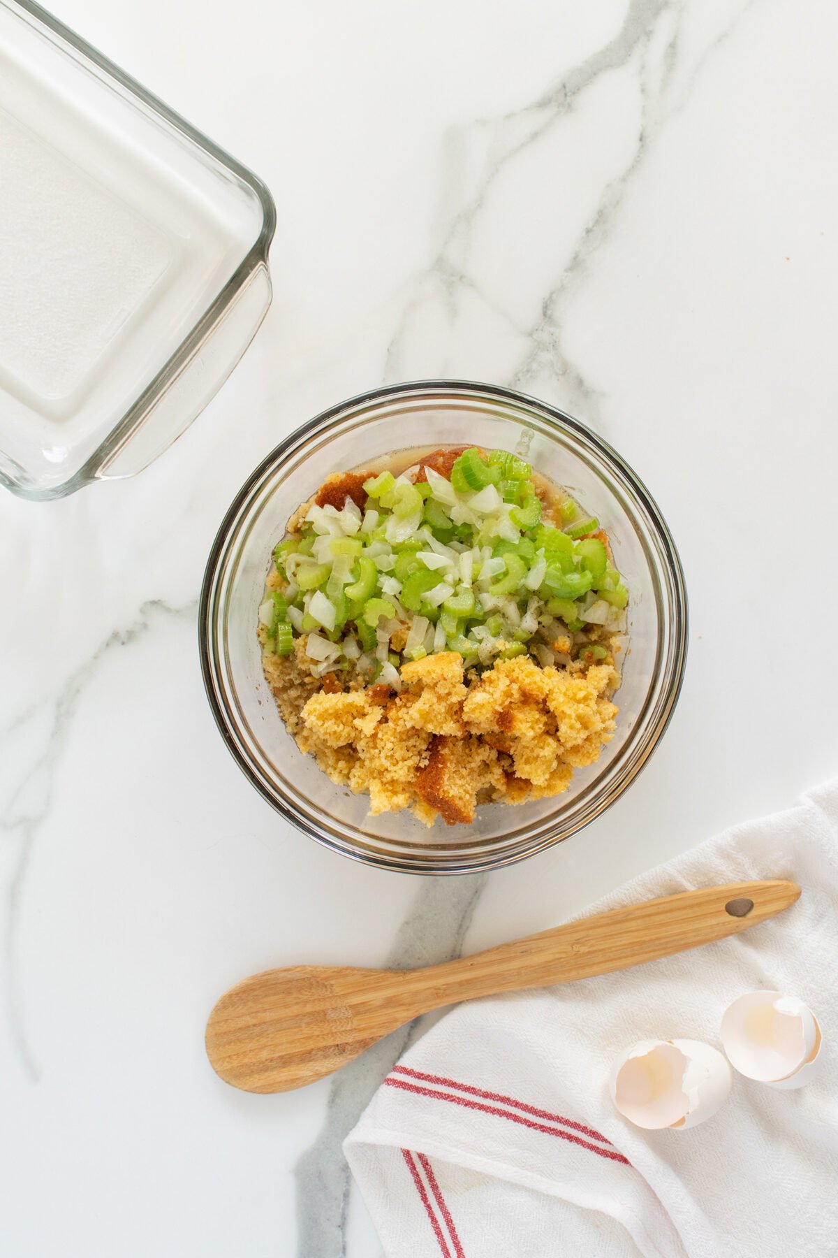 A glass bowl with chopped celery, onions, and crumbled cornbread mixture on a marble countertop; beside it are a wooden spoon, broken eggshells, a red-striped towel, and an empty glass baking dish.
