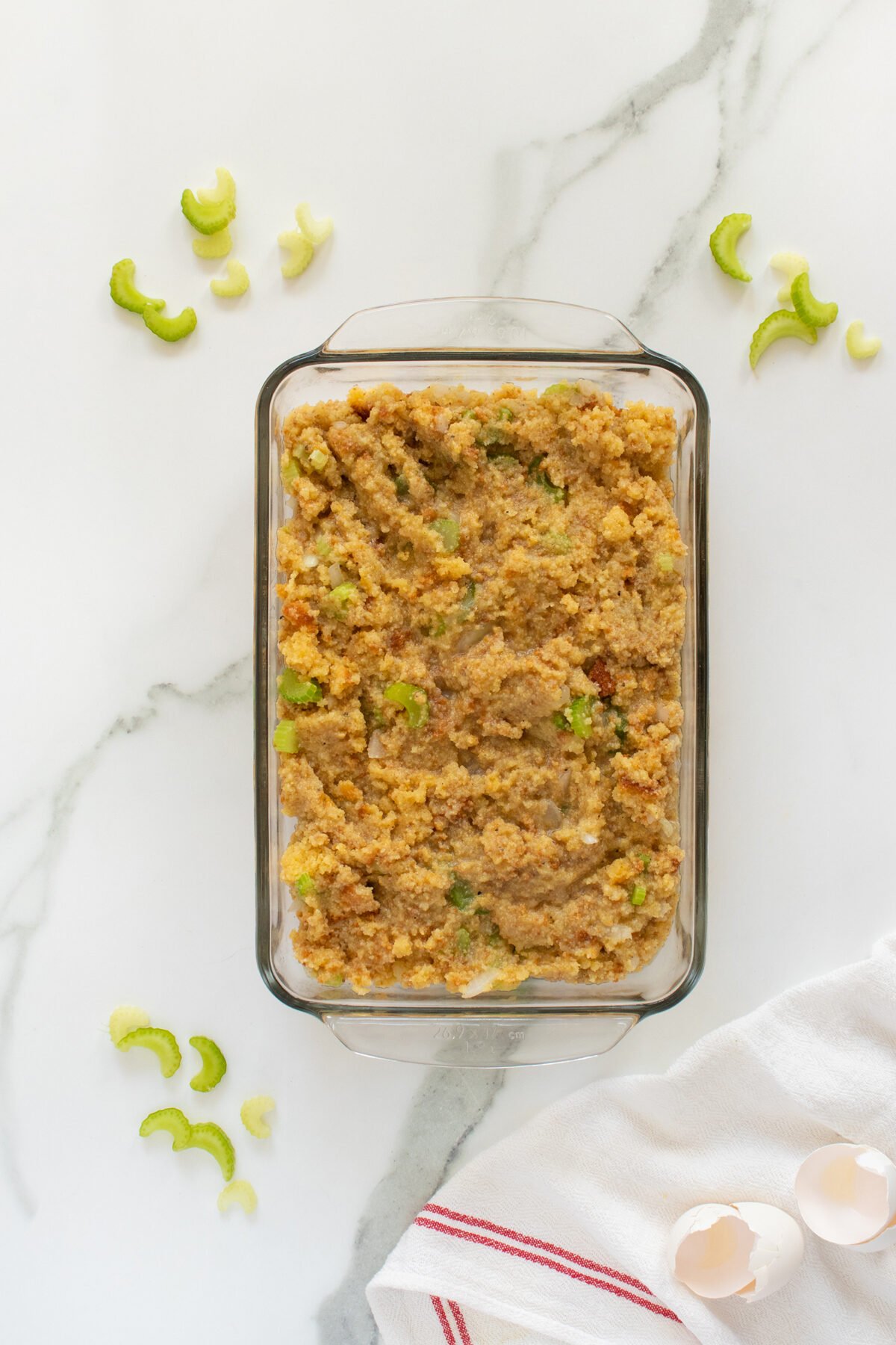A glass baking dish filled with cornbread stuffing on a marble surface, surrounded by sliced celery pieces, eggshells, and a white towel with red stripes.