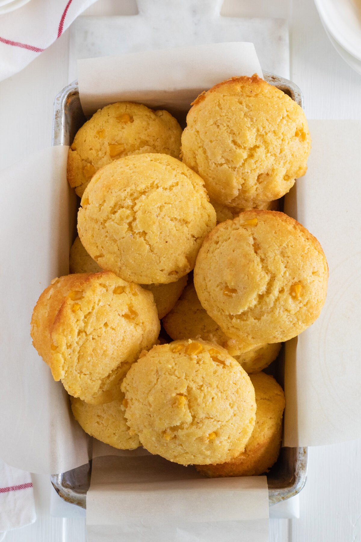 A rectangular metal baking dish lined with parchment paper holds several golden brown cornbread muffins, arranged closely together on a white surface.