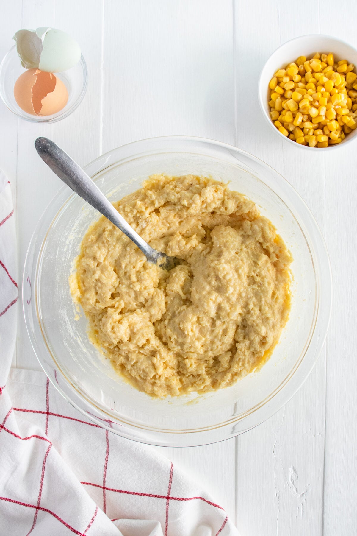 A glass bowl filled with cornbread batter and a spoon sits on a white table. Nearby are cracked eggshells, a bowl of corn kernels, and a white cloth with red stripes.