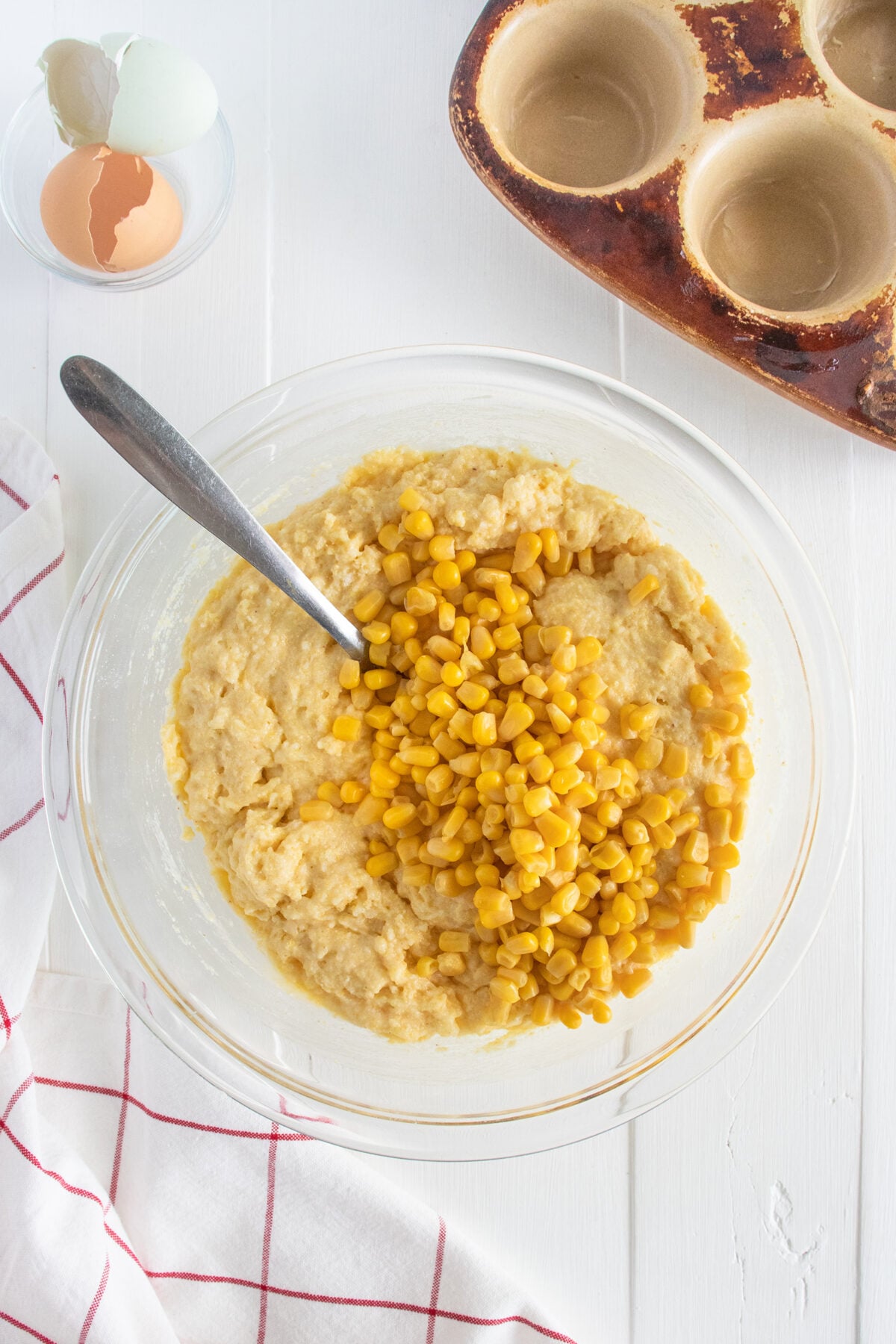 A glass bowl with cornbread batter and corn kernels, a spoon inside, sits on a white table next to cracked eggshells, an empty muffin tin, and a red-striped kitchen towel.