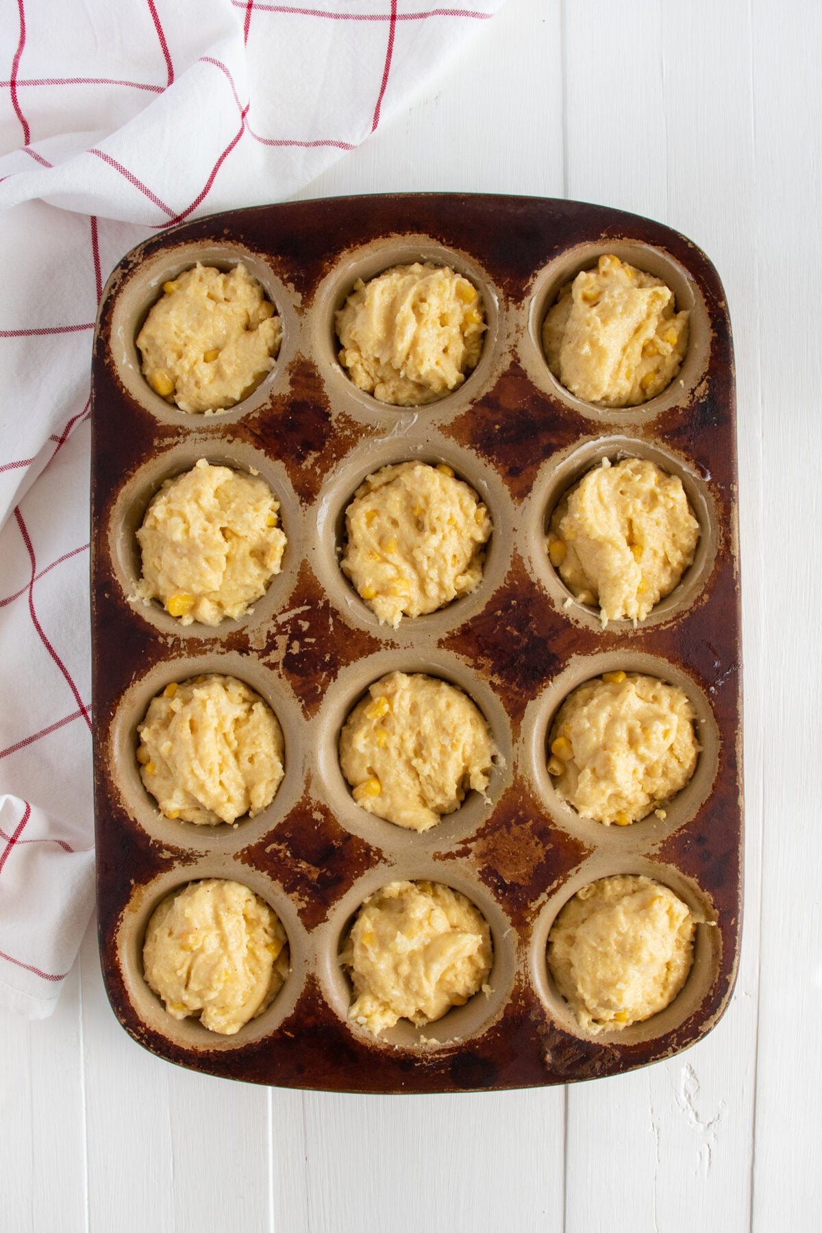 A muffin tin filled with raw corn muffin batter, ready to be baked, sits on a white wooden surface next to a white and red checkered kitchen towel.