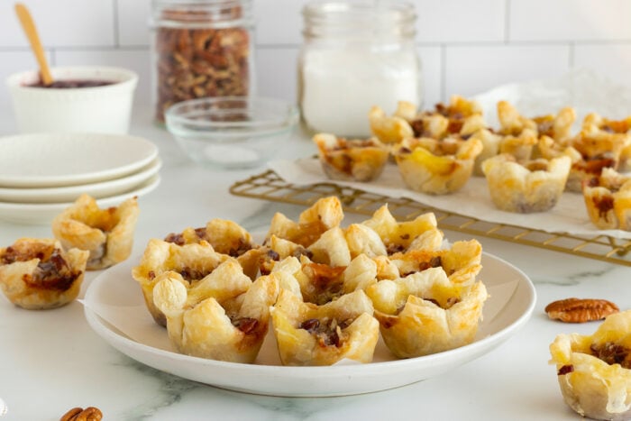 A plate of mini pecan tarts sits on a white countertop, with more tarts cooling on a wire rack in the background. Glass jars, small bowls, and a stack of plates are also visible.