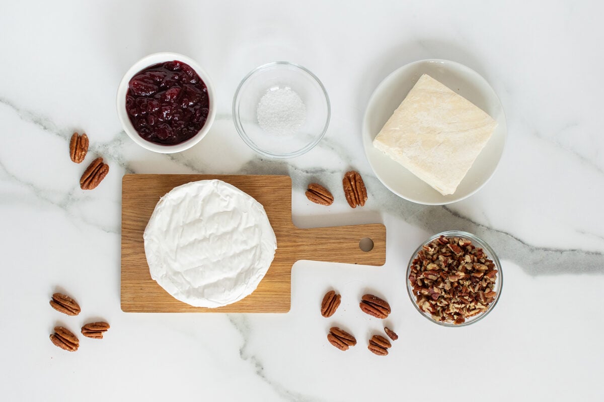 A round wheel of brie cheese on a wooden board, surrounded by a block of parmesan, a bowl of cherry jam, a bowl of chopped pecans, a bowl of salt, and scattered whole pecans on a white marble surface.