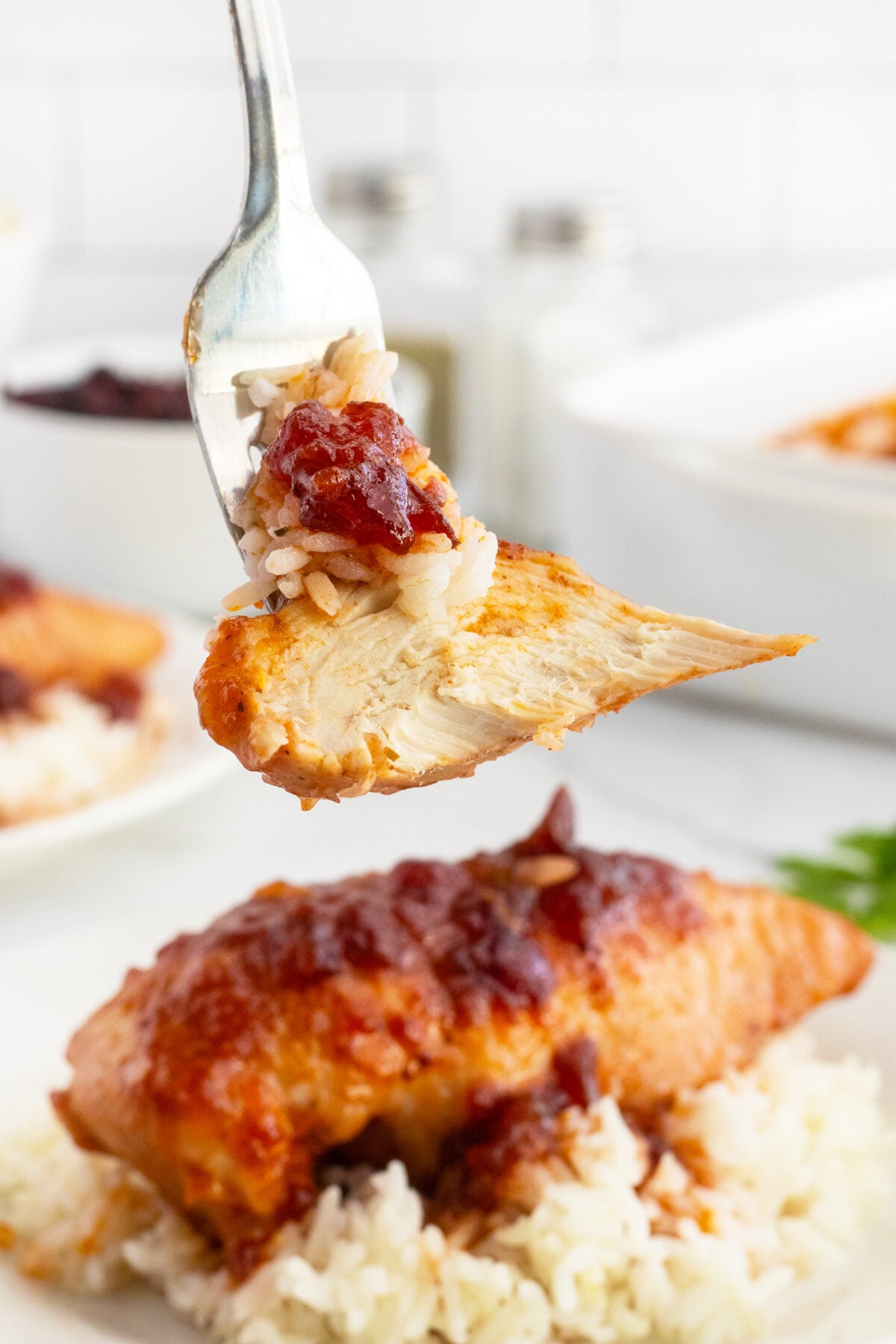 A fork holds a piece of baked chicken with rice and sauce, above a plate of white rice topped with more saucy chicken. The background shows additional servings and a white, bright kitchen setting.
