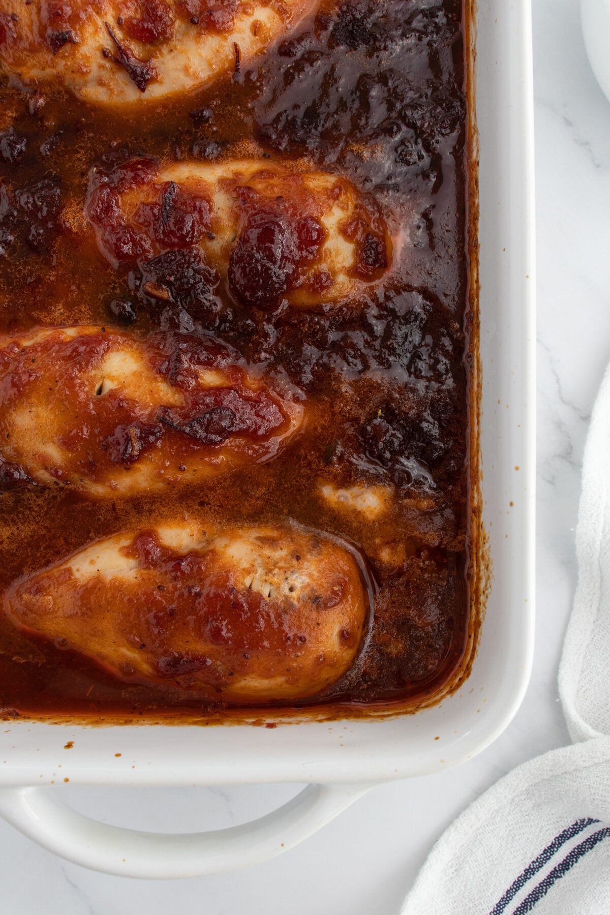 Four baked chicken breasts in a white casserole dish, covered in a thick, glossy, dark red sauce. The dish is set on a white marble surface with a white towel visible at the edge.