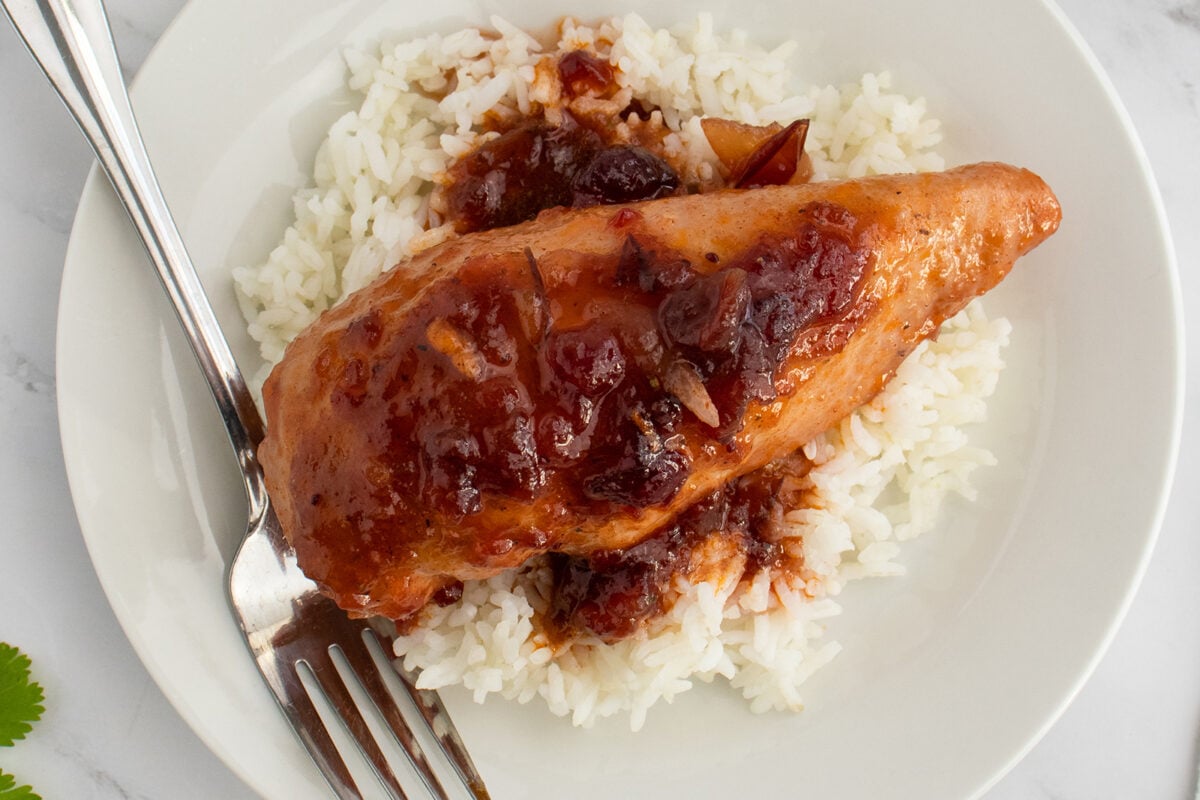 A cooked chicken breast topped with a reddish sauce sits on a bed of white rice on a white plate, with a fork resting on the side.