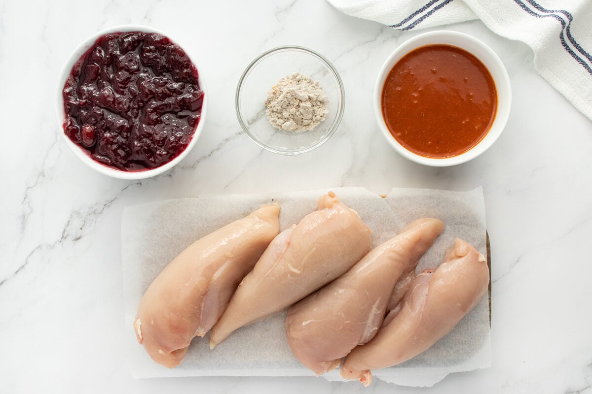 Four raw chicken breasts on parchment paper, next to bowls containing cranberry sauce, powdered seasoning, and an orange-brown sauce, all arranged on a white marble surface.