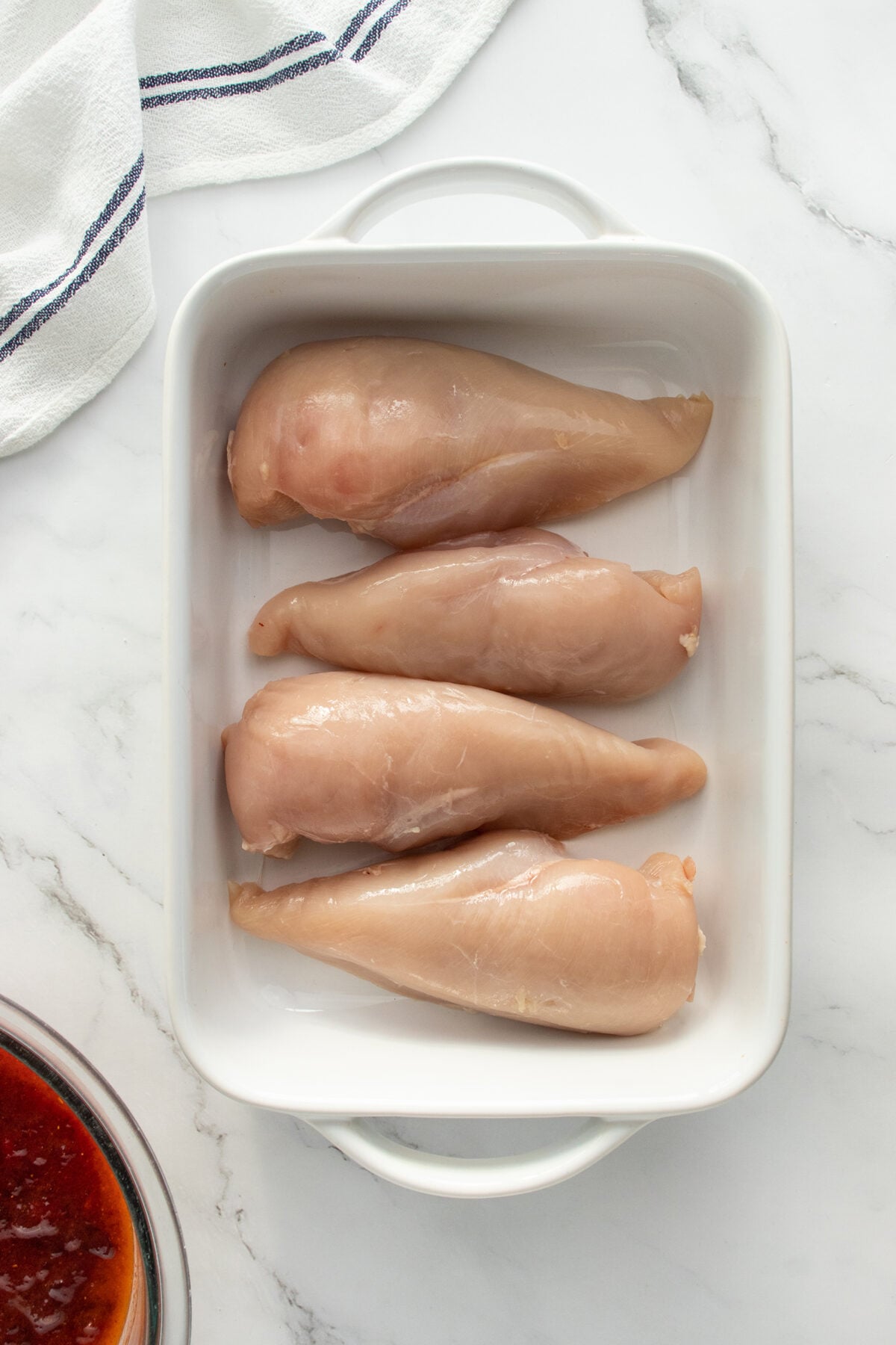 Four raw chicken breasts are placed side by side in a white rectangular baking dish on a marble countertop, with a striped towel and a bowl of red sauce partially visible.