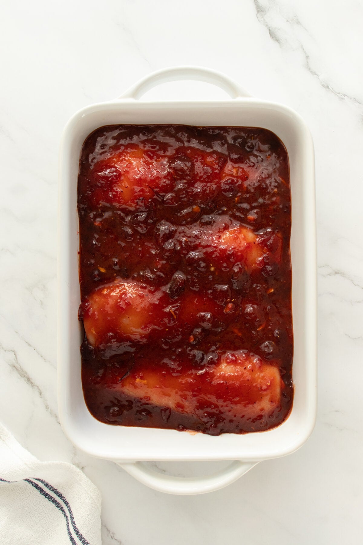A white baking dish on a marble surface contains three raw chicken breasts covered in a thick, red, fruit-based sauce. A white towel with a dark stripe is partially visible in the corner.