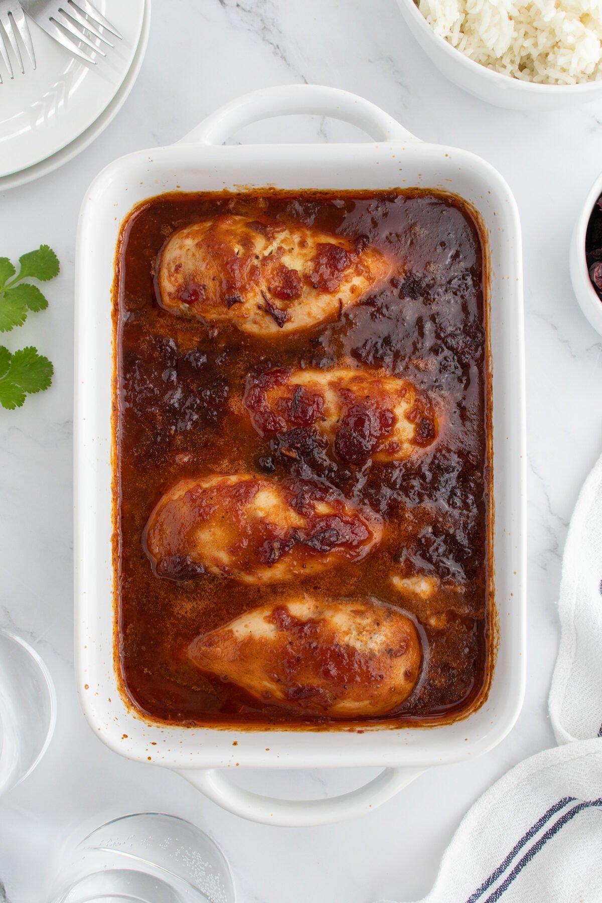 Four baked chicken breasts in a rectangular white dish covered in a rich brown sauce, surrounded by plates, utensils, a bowl of white rice, and a sprig of cilantro on a white surface.