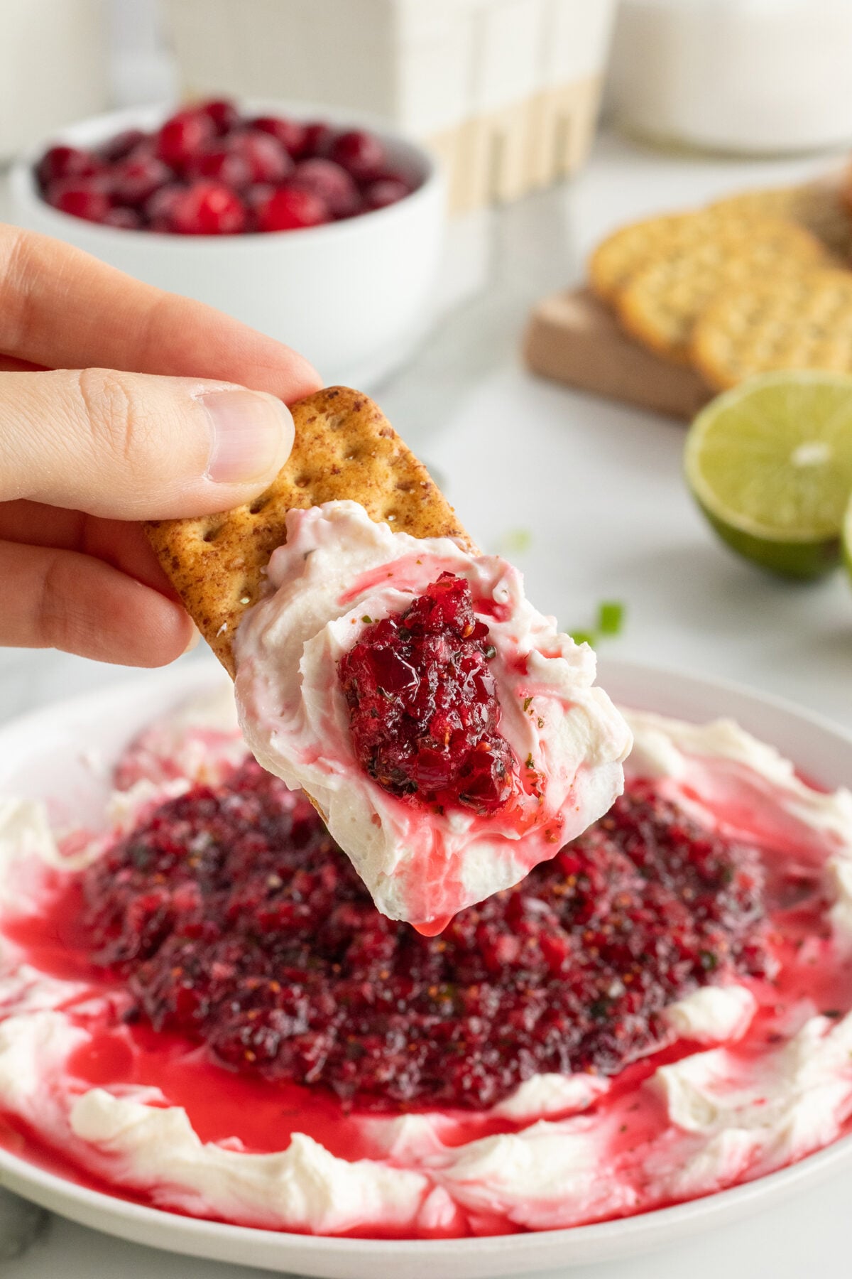 A hand holds a cracker topped with creamy cheese and cranberry sauce, above a plate of cheese spread with cranberry sauce. Crackers, a bowl of cranberries, and a sliced lime are visible in the background.