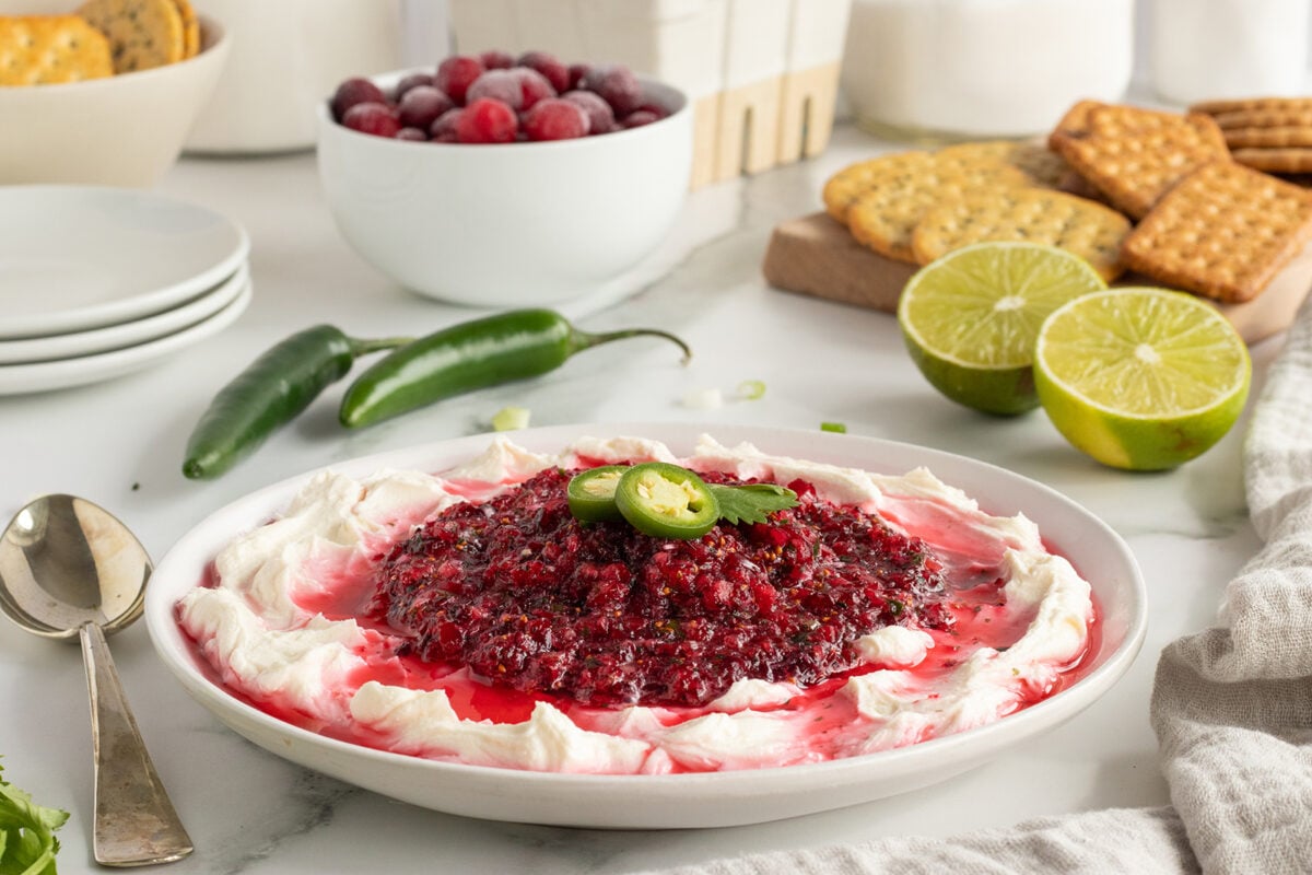 A plate of creamy dip topped with cranberry salsa and sliced jalapeños, surrounded by lime halves, crackers, green chili peppers, and a bowl of fresh cranberries on a white countertop.