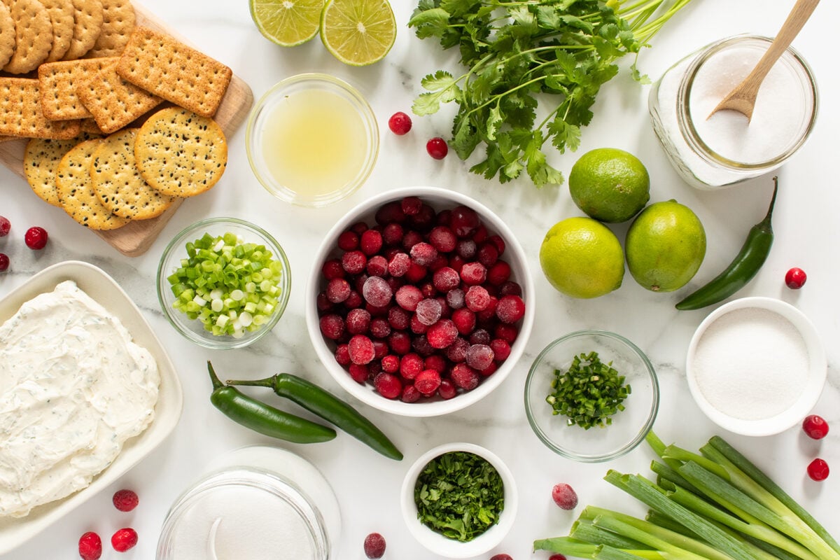 An assortment of ingredients on a white surface, including a bowl of cranberries, green onions, limes, cilantro, jalapeños, round and square crackers, cream cheese, lime juice, sugar, and salt.