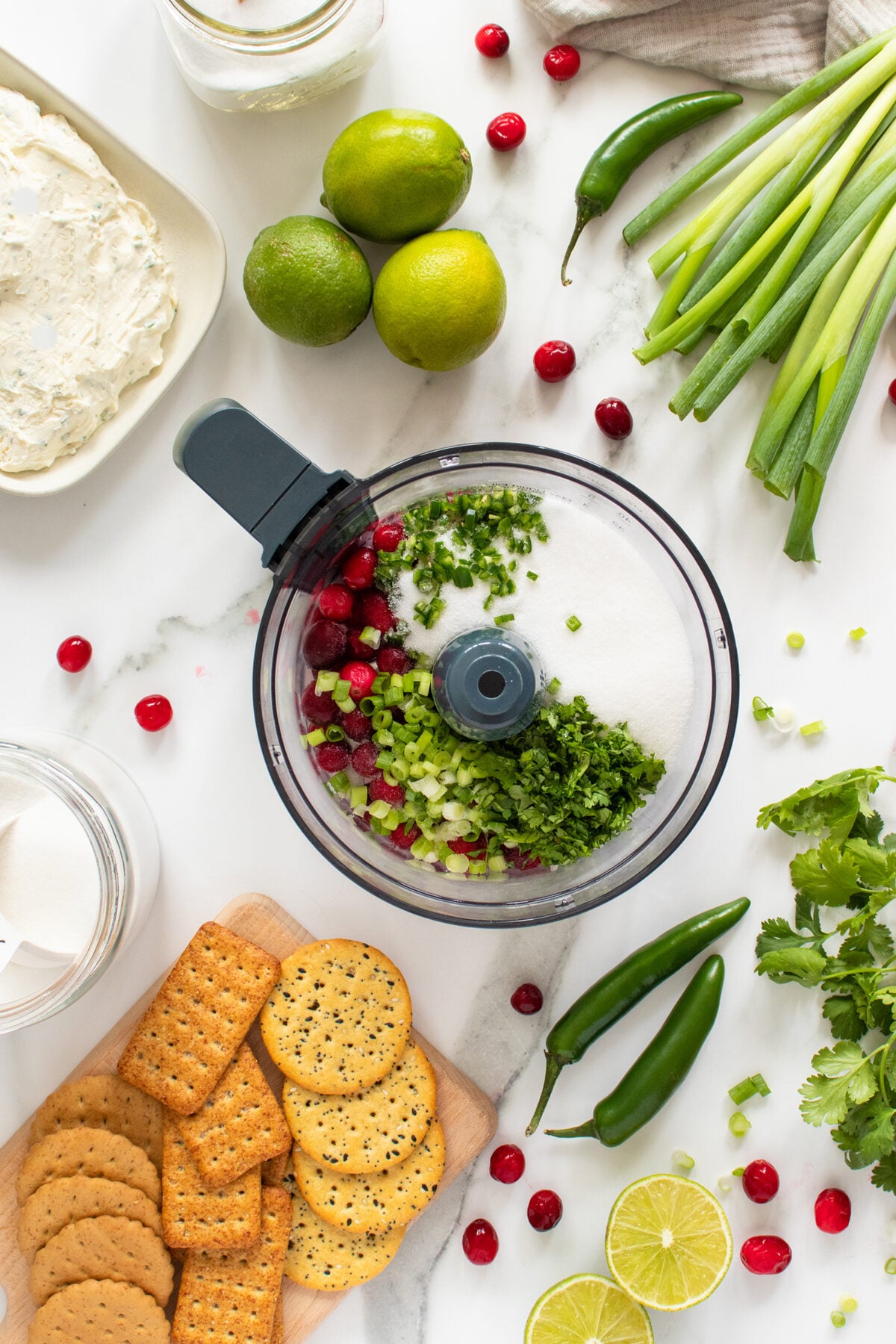 A food processor with cranberries, chopped green onions, cilantro, and sugar sits on a white surface surrounded by crackers, limes, jalapeños, cream cheese, cilantro, and green onions.