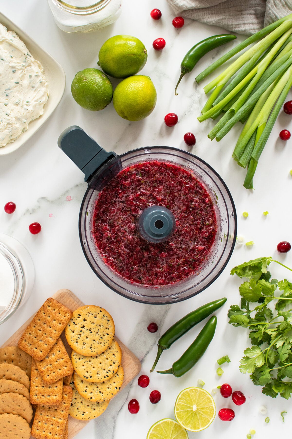 A food processor with blended cranberry mixture is surrounded by crackers, limes, green onions, cilantro, whole cranberries, jalapeños, lime slices, and a jar of spread, all arranged on a white countertop.