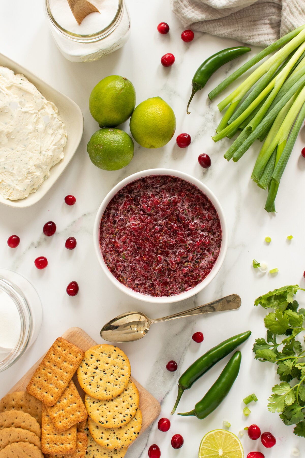 A bowl of cranberry salsa surrounded by crackers, cream cheese, limes, green onions, cilantro, jalapeños, and scattered cranberries on a white marble surface.
