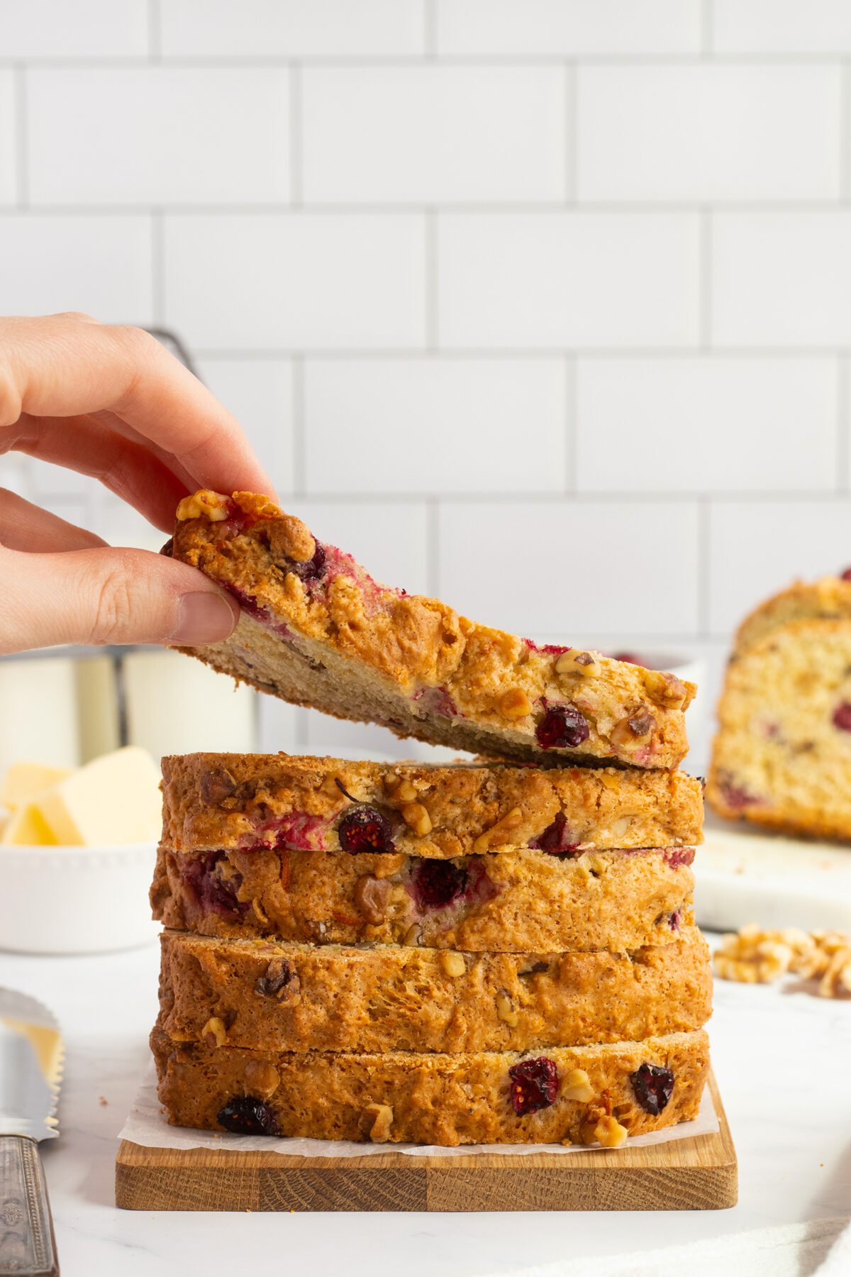 A hand lifts a slice from a stack of sliced nut and berry bread on a wooden board. Butter, walnuts, and more bread are visible in the background against a white tiled wall.