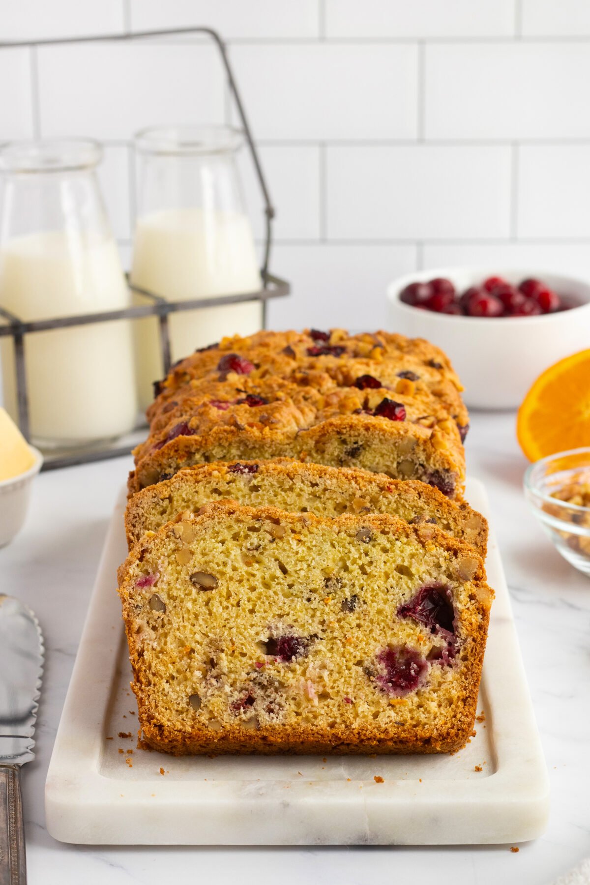 Sliced cranberry nut bread on a white marble tray, with glasses of milk, a bowl of cranberries, an orange half, and chopped nuts in the background on a white countertop.