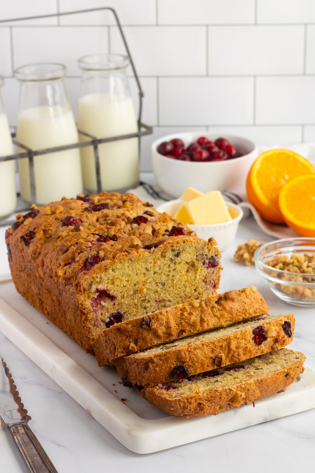 A loaf of cranberry orange bread, partially sliced, sits on a marble board. Nearby are orange halves, a bowl of cranberries, butter, walnuts, and glass bottles of milk against a white tiled background.