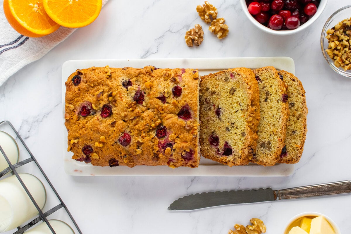 A loaf of cranberry walnut bread on a white plate, with three slices cut. Surrounding it are orange slices, cranberries, walnuts, eggs, a knife, and a small bowl of butter on a marble surface.