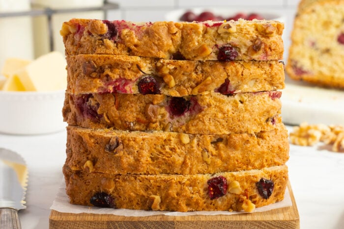 Five thick slices of cranberry nut bread are stacked neatly on a wooden board. The bread is golden brown with visible pieces of cranberry and walnuts throughout. A knife and butter are in the background.