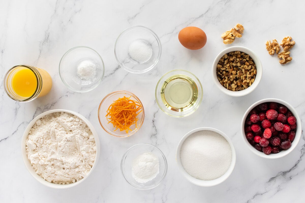 A flat lay of baking ingredients on a marble surface, including flour, orange juice, orange zest, sugar, oil, an egg, salt, baking powder, baking soda, walnuts, and fresh cranberries in bowls.