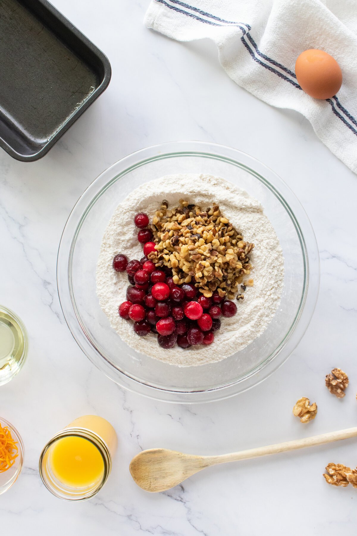A glass bowl with flour, cranberries, and chopped walnuts sits on a white countertop, surrounded by an egg, orange juice, oil, a baking pan, orange zest, a wooden spoon, and a towel.