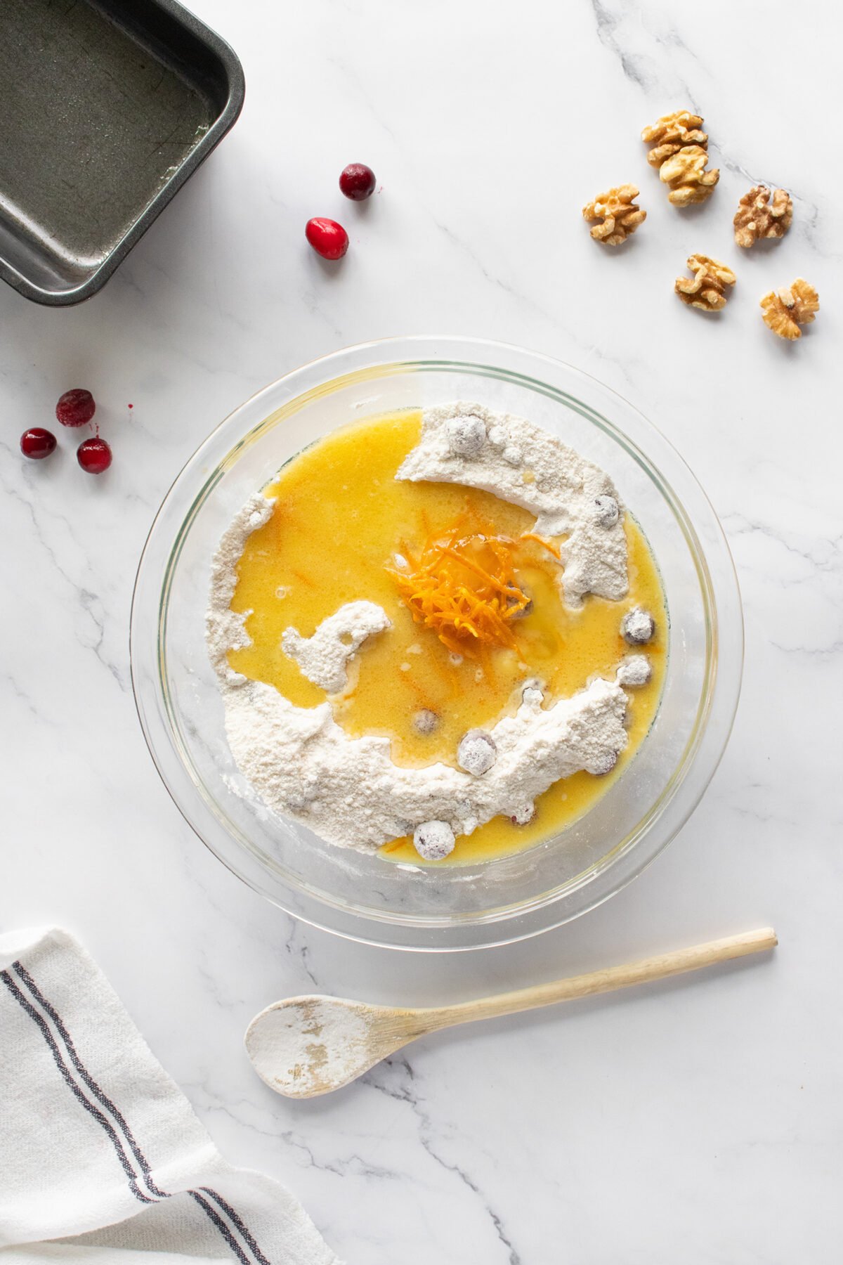 A glass bowl with flour, melted butter, cranberries, and orange zest sits on a marble surface. Nearby are walnuts, cranberries, a metal pan, a wooden spoon, and a white towel with black stripes.