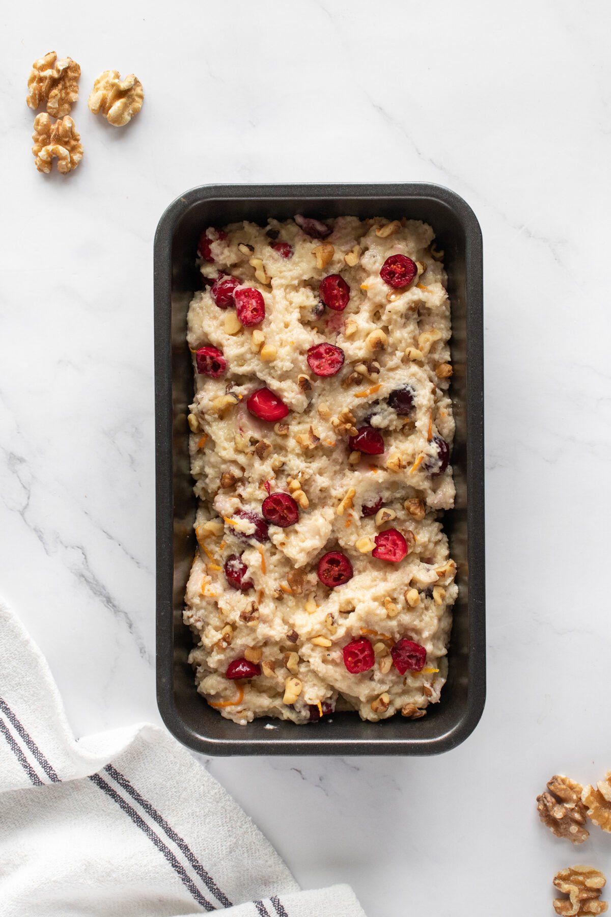 A loaf pan filled with unbaked bread batter topped with cranberries and walnuts sits on a white marble surface. Two walnut halves and a white towel with gray stripes are nearby.