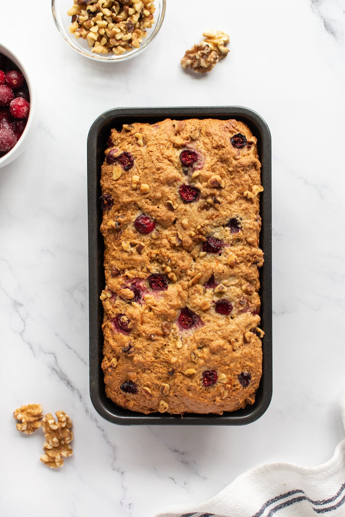 A loaf of cranberry walnut bread in a rectangular pan, topped with visible cranberries and walnut pieces, sits on a white marble surface next to bowls of walnuts and cranberries.