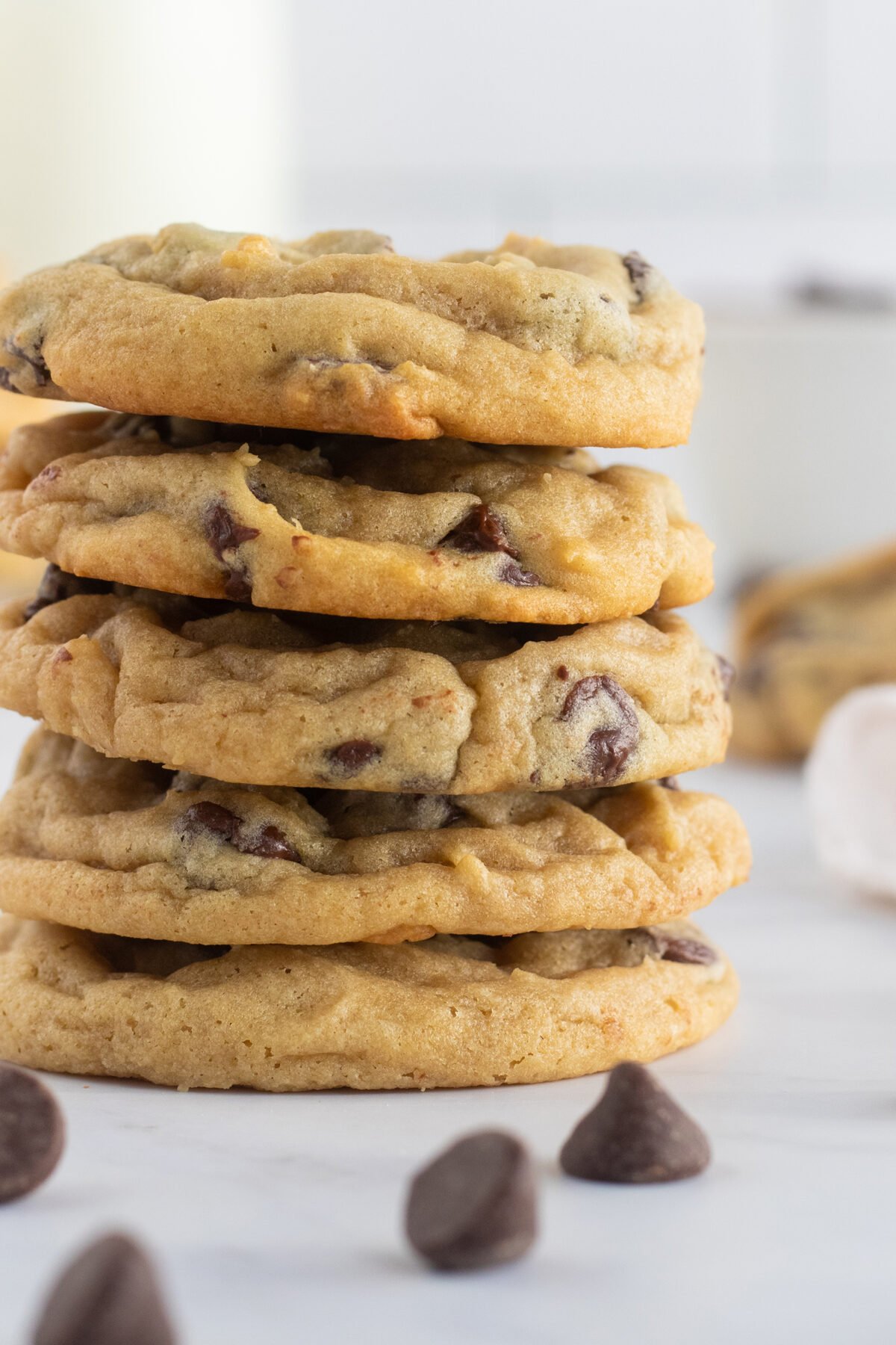 A stack of five thick, golden chocolate chip cookies sits on a white surface with a few chocolate chips scattered nearby. The cookies look soft and chewy, with chocolate chips visible throughout.