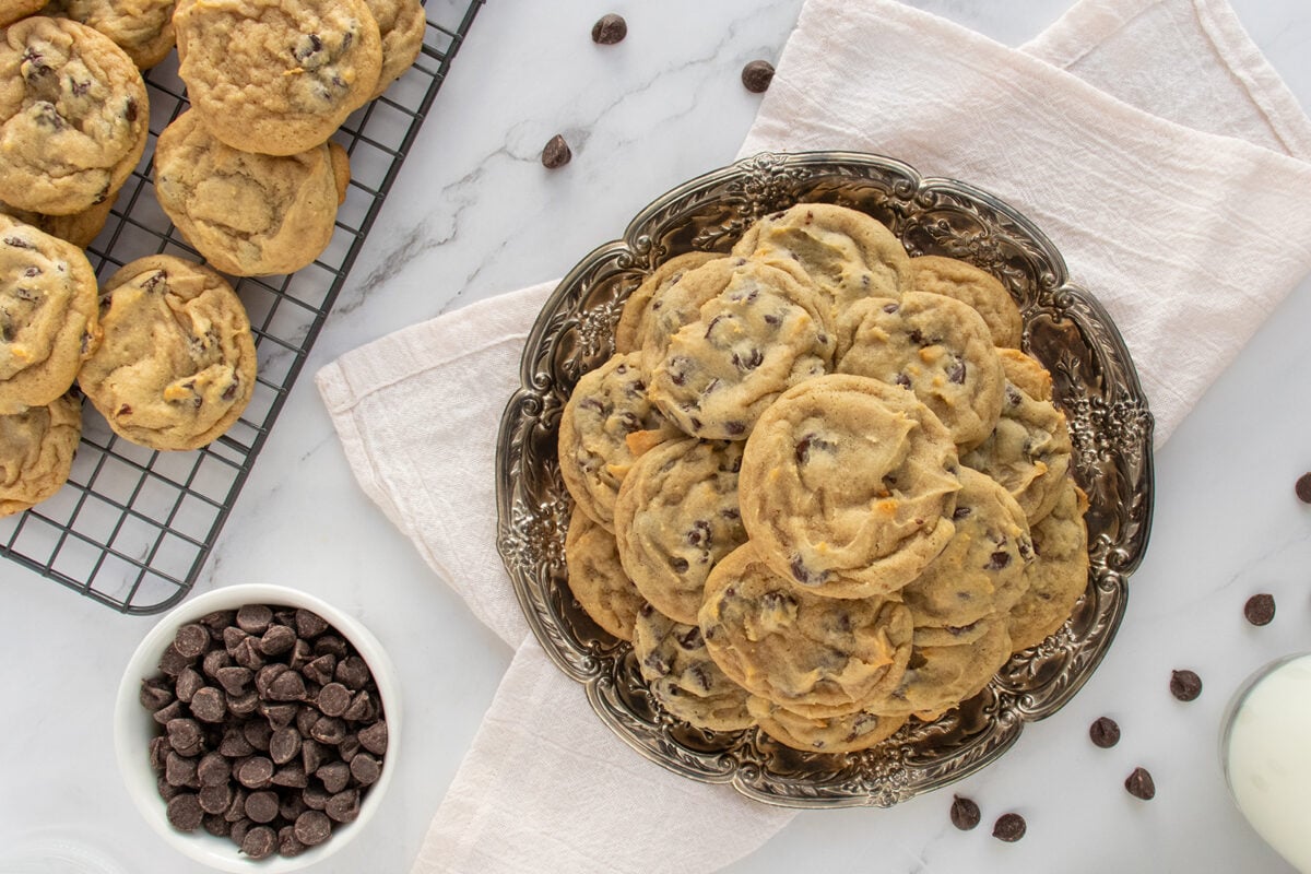 A plate of chocolate chip cookies sits on a cloth napkin, with more cookies on a cooling rack nearby. A small bowl of chocolate chips and a glass of milk are also on the marble countertop.