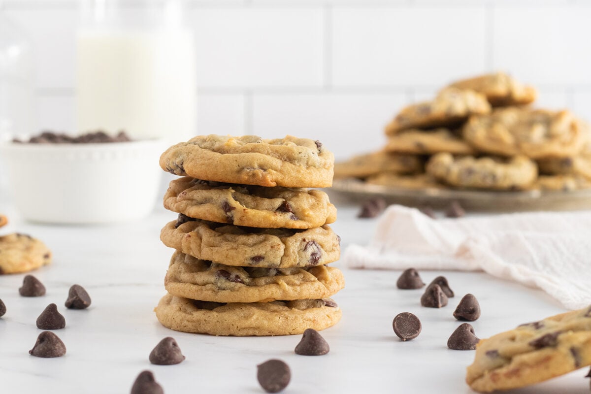 A stack of chocolate chip cookies on a white surface, surrounded by scattered chocolate chips, with more cookies and a glass of milk in the background.
