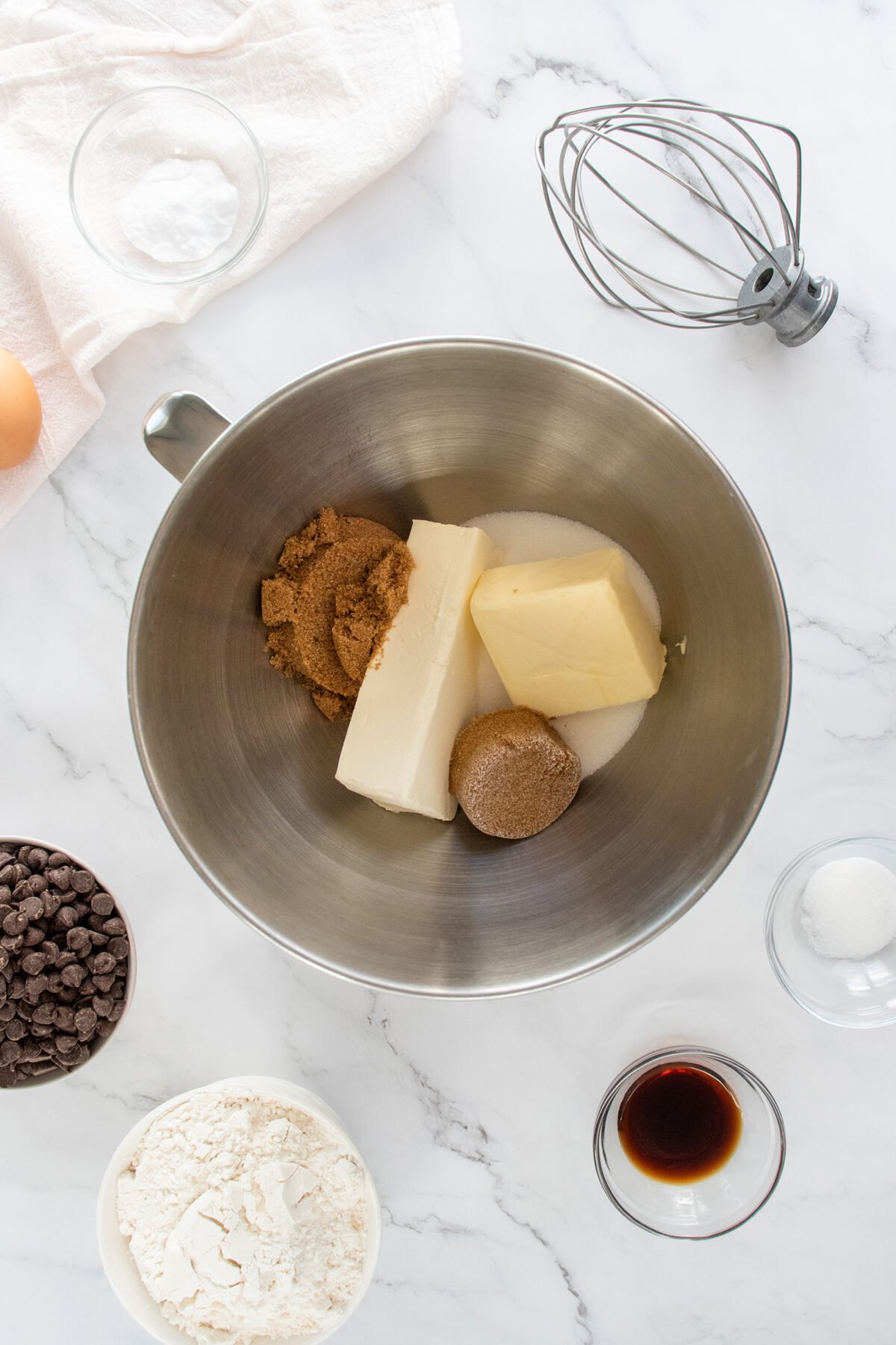 A mixing bowl with butter, brown sugar, and white sugar, surrounded by a bowl of flour, chocolate chips, vanilla extract, an egg, salt, baking soda, and a whisk on a white marble surface.