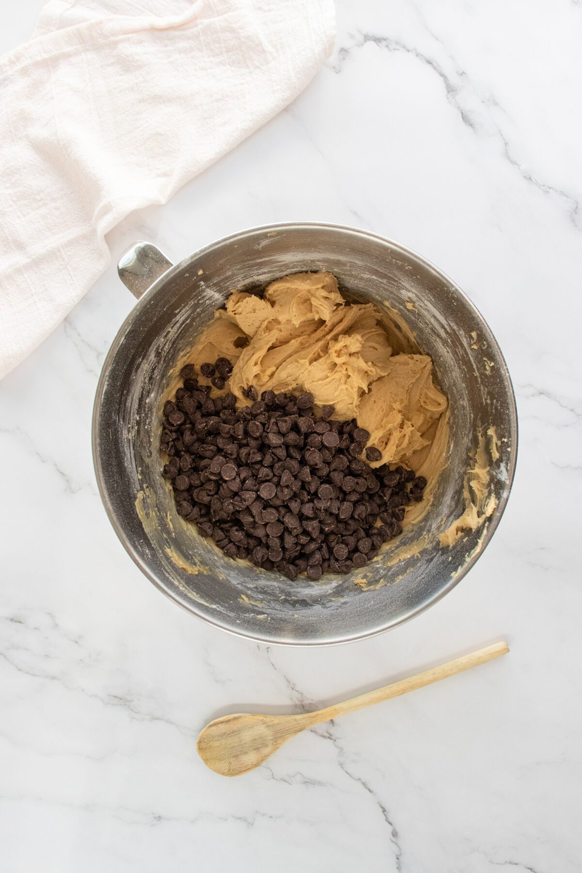 A metal mixing bowl with cookie dough and chocolate chips sits on a marble surface. Nearby are a wooden spoon and a light cloth napkin.