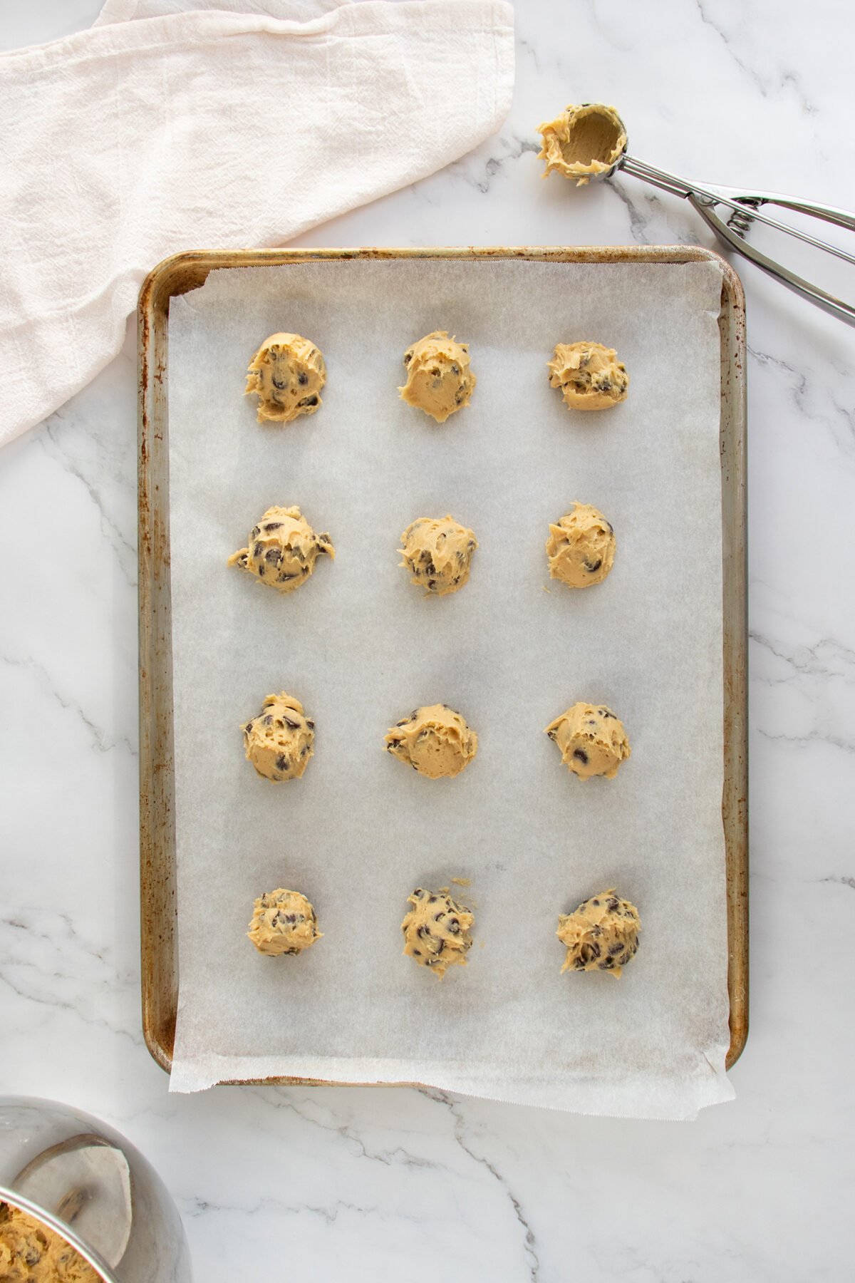 A baking tray lined with parchment paper holds twelve scoops of chocolate chip cookie dough, ready to bake. Nearby are a metal cookie scoop with dough and a white cloth on a marble countertop.