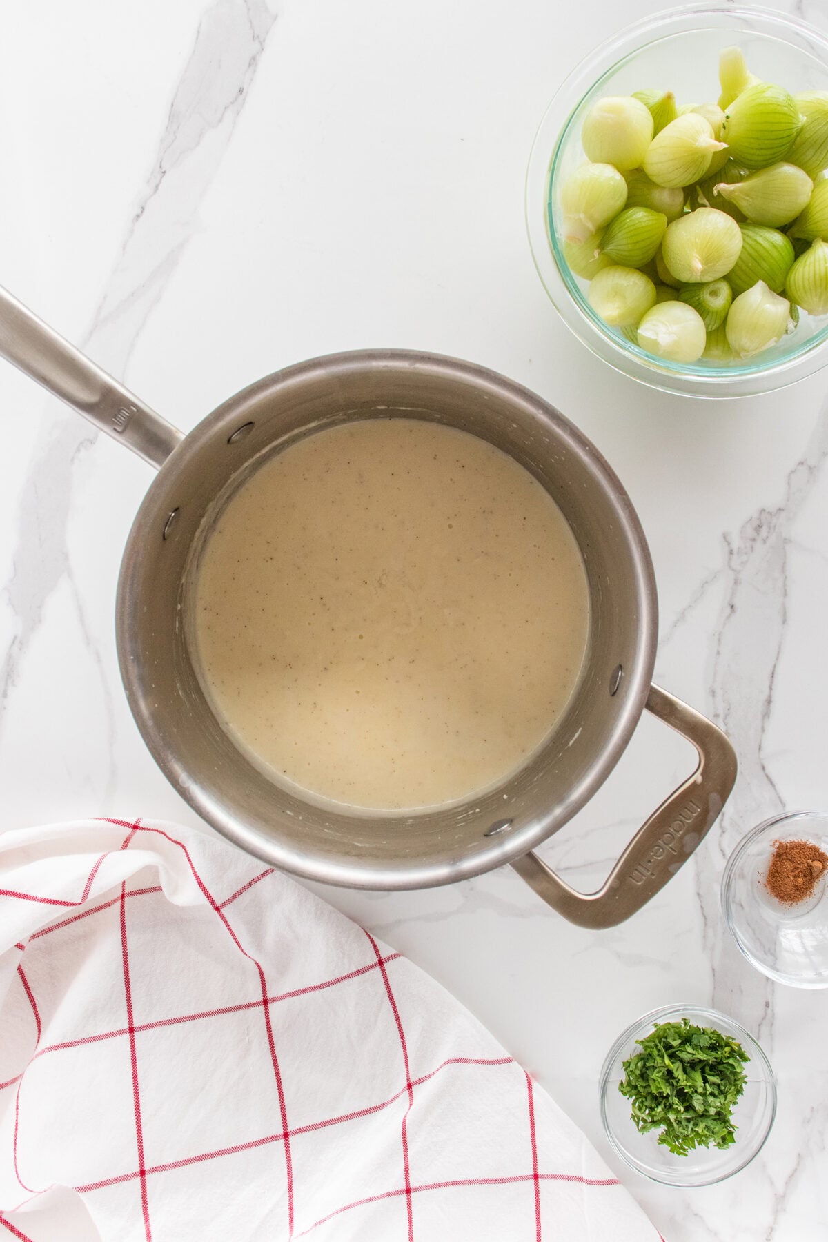 A saucepan of creamy white sauce on a marble countertop, next to a bowl of peeled green tomatillos, a small bowl of chopped herbs, a small amount of ground spice, and a white towel with red lines.