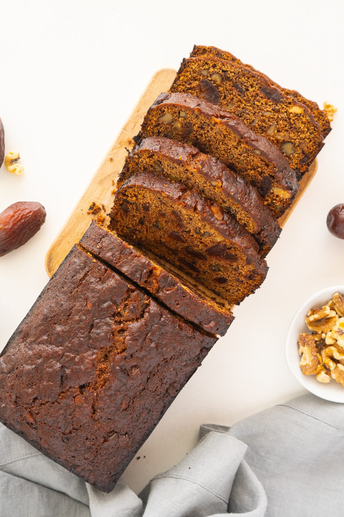 A loaf of date and walnut bread, partially sliced on a wooden board. Slices show pieces of dates and walnuts. Whole dates, walnut pieces, and a gray cloth are arranged around the bread.