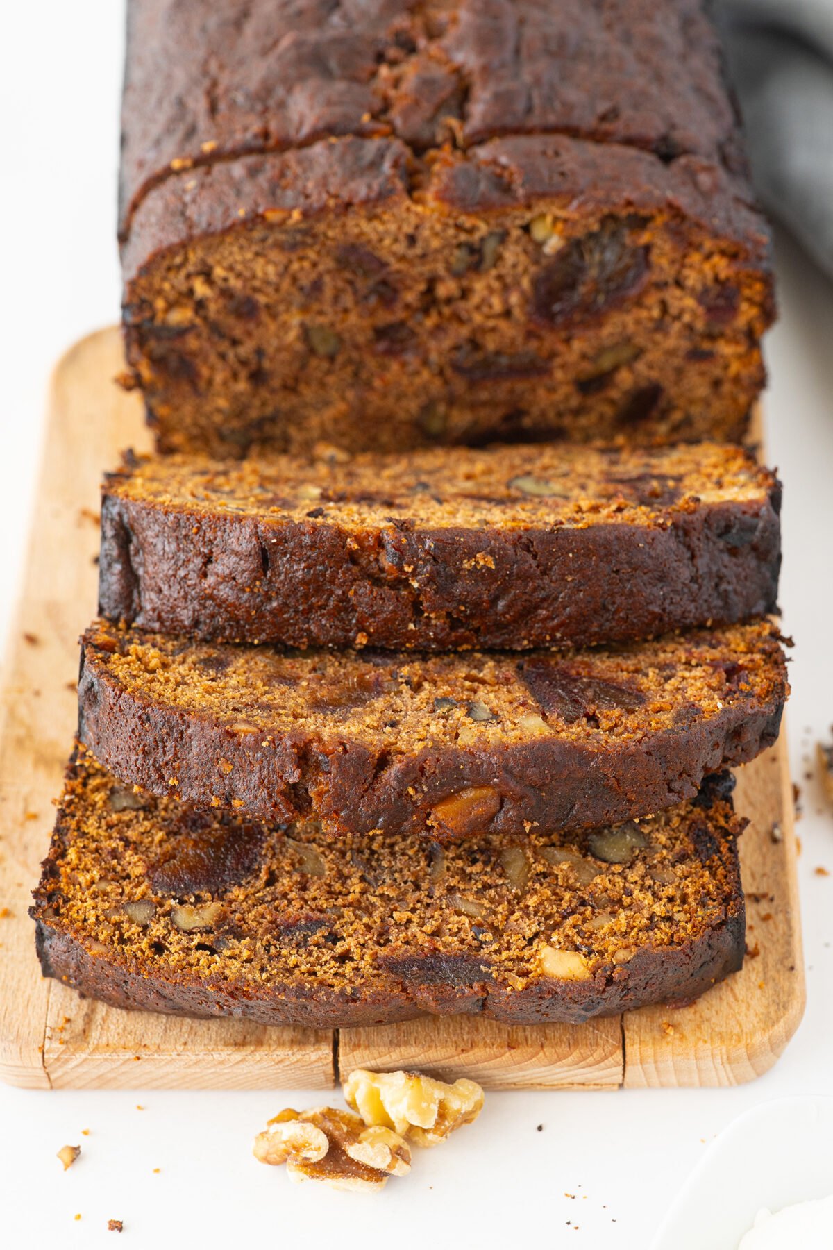 A sliced loaf of dark, moist banana bread with visible nuts and pieces of fruit, arranged on a wooden cutting board. A few walnut pieces are scattered nearby.