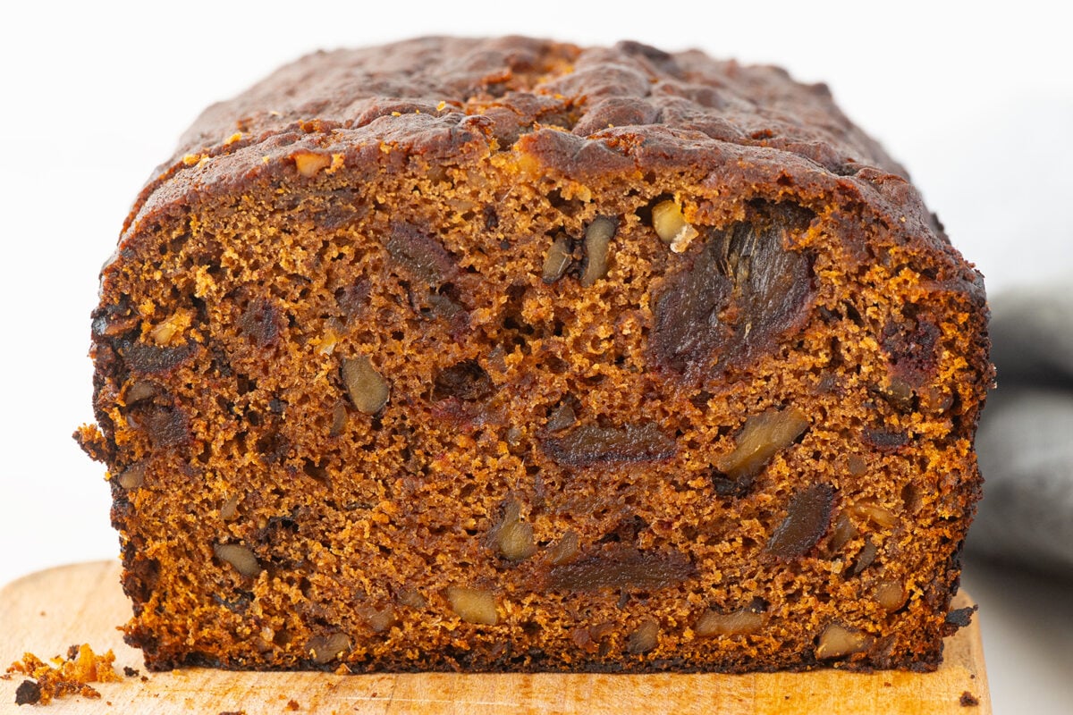 A close-up of a sliced loaf of fruit and nut bread, showing a dense, moist texture with visible pieces of nuts and dried fruit throughout. The exterior is dark brown with a slightly uneven crust.