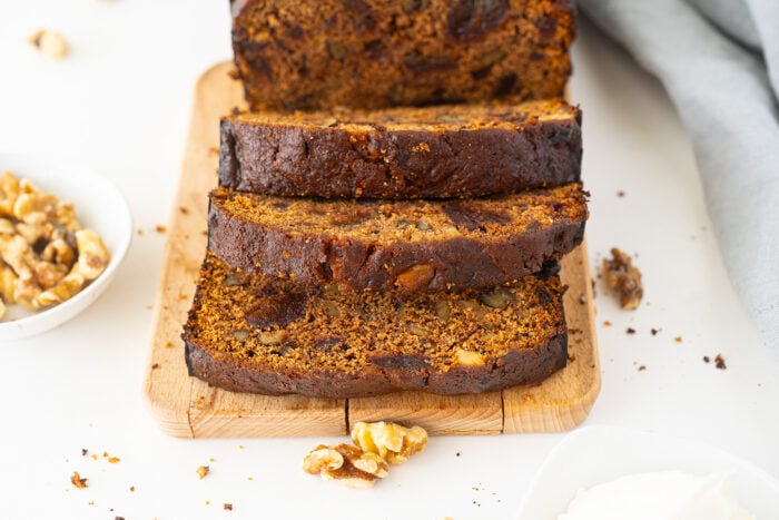 Three slices of dark, moist banana bread with nuts are stacked on a wooden cutting board. There are some walnuts scattered around and a small white bowl of walnuts beside the bread.