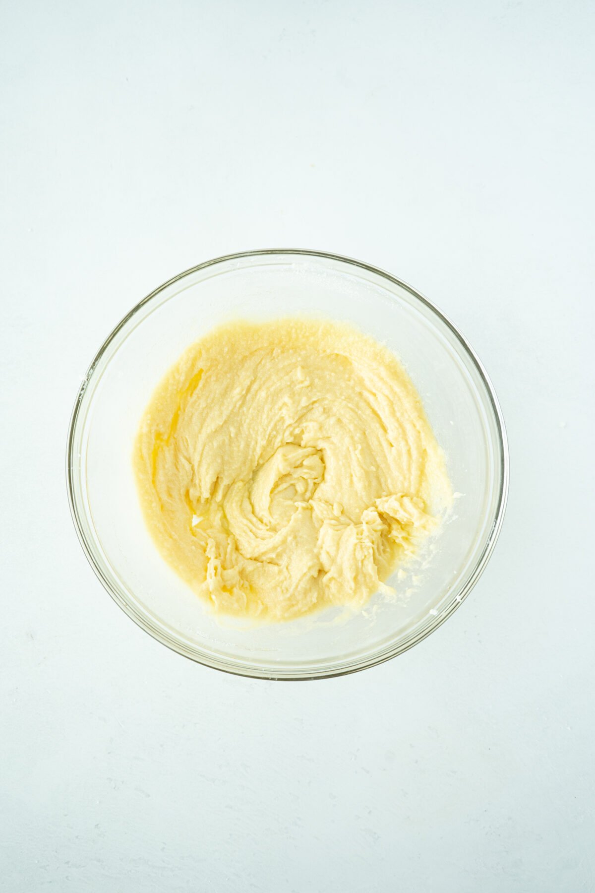 A clear glass bowl filled with creamy, pale yellow cake batter sits on a white surface, viewed from above.